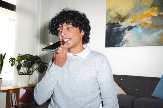 An Arab young man is using the voice command on his smartphone in a modern living room. He is wearing a casual sweater and standing near a sofa and plants.