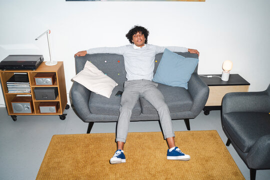 An Arab young man is relaxing on a gray sofa in a modern living room. He is smiling and has his arms outstretched on the back of the couch.