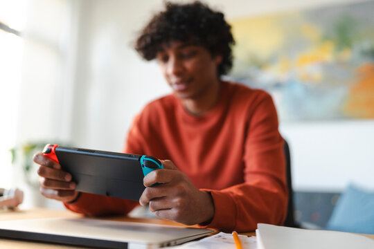 An Arab young man is playing a handheld video game console at a desk. He is taking a break from studying or working.