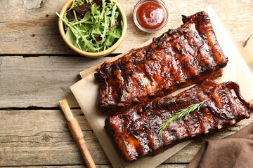 Tasty roasted pork ribs served with rosemary, sauce and salad on wooden table, flat lay