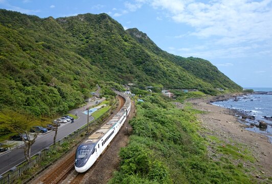 A sleek train glides along the coast of Shicheng, Yilan, between lush green hills and the rocky sea. The blue sky, ocean, and winding tracks create a peaceful, scenic blend of nature and travel.