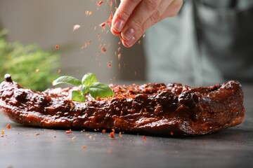 Woman sprinkling spices onto ribs at grey table indoors, closeup