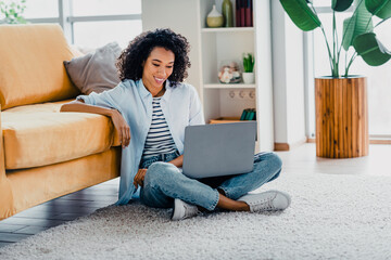 Young woman with curly hair relaxing at home, sitting on a carpet using a laptop in a bright, cozy...