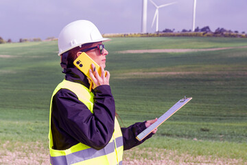 Female engineer using mobile phone and holding clipboard in wind turbine field