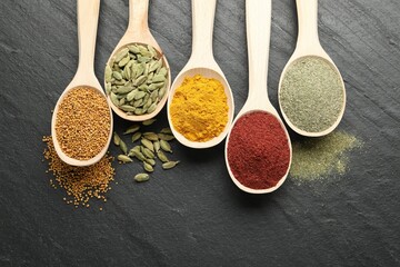 Different aromatic spices in spoons on black table, flat lay