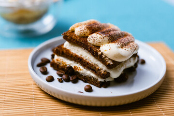 Porción de tiramisú con capas visibles de bizcocho y crema espolvoreada con cacao, decorado con granos de café enteros. fondo colorido y mantel bambú. Postre italiano.