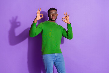 Young man wearing a green sweatshirt smiling and showing perfect okay gestures against a vibrant...