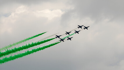 Fighter jets perform an aerial display, leaving bold green and white smoke trails in the sky, symbolizing the Pakistan flag. The jets fly in a tight formation against a soft sky backdrop.
