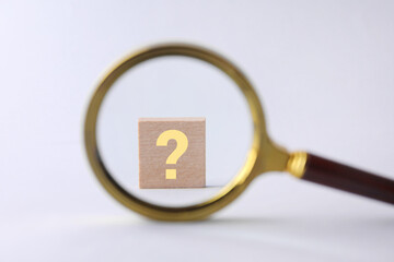 Wooden cube with question mark on light grey background, view from magnifying glass