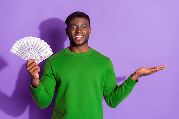 Young man in a green sweater holding cash in hand on a purple background showcasing a cheerful and...