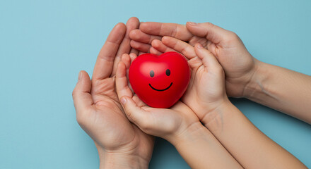 A red heart with a smiley face held in two sets of hands on a light blue background studio shot