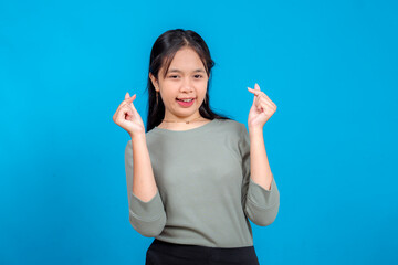 Fototapeta premium Happy young Asian girl smiling and making double finger heart gestures. Her bright expression, casual outfit, and playful charm pop against a clean blue studio background.