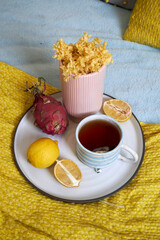 A tray with tea, flowers, and fruit placed on a bed with bright yellow, pink, blue, and gray...