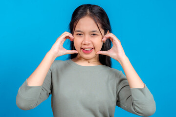 Naklejka premium Cheerful young Asian woman with long hair smiling and playfully framing her cheeks with her hands against a blue background. Joyful facial expression for lifestyle and advertising use.