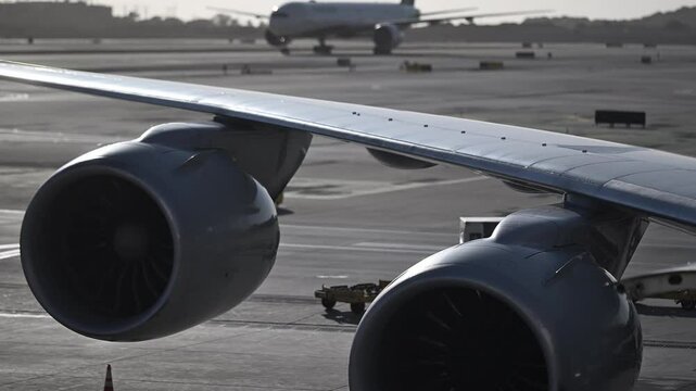 Detailed view of large twin jet engines and wing of a commercial aircraft on the airport tarmac. Another airplane is seen taxiing in the distance during early morning or evening light.