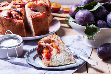Plum pie with filling and sugar crust, sprinkled with powdered sugar and plums on a wooden table.
