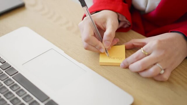 A woman wearing a red jacket writes "Meetings" on a sticky note. She's using a pen and has a laptop nearby.  She's likely making a to-do list or jotting down reminders for upcoming meetings.