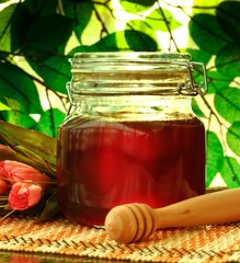 This image showcases a clear glass jar filled with golden honey, complimented by a wooden honey dipper and surrounded by vibrant flowers, set against a leafy backdrop. - obrazy, fototapety, plakaty