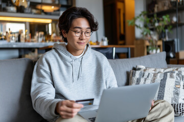Obraz premium A young Chinese Asian man sitting on the sofa at home in a living room with a laptop on the lap and holding a credit card. Online shopping, payment, purchase, buy. E-commerce.