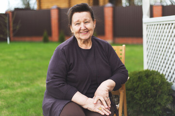 Smiling elderly woman around 70 80 years old sitting on a wooden chair in green backyard, wearing dark clothes and looking at camera, concept aging, happiness, outdoor lifestyle