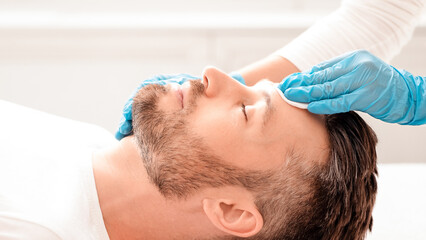 Side view of unrecognizable cosmetologist cleansing bearded man face with toner before or after treatment, using cotton pad. Middle-aged businessman having beauty procedures at spa