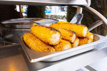 Mouthwatering grilled corn cobs stacked in a silver tray. The golden kernels show grill marks. A blurred background suggests an outdoor event or food vendor