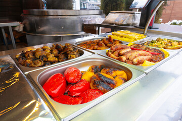 Close-up of different types of grilled food, including vegetables and meat, presented in stainless steel containers at an outdoor vendor. Vibrant colors and textures are highlighted