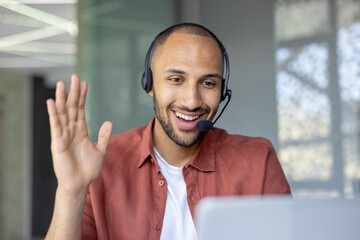 A smiling man wearing a headset waves during a video call, engaging in a friendly and professional...