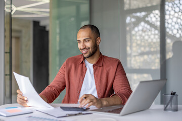 A smiling man in a red shirt reviews documents and uses a calculator at his desk in a modern office setting. A laptop sits beside him.
