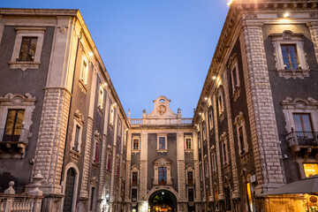 Fototapeta premium Illuminated Uzeda Gate between historic buildings in the Cathedral Square of Catania, Sicily, Italy, at dusk