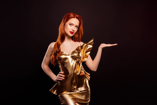 Stunning red haired woman in a luxurious golden dress posing gracefully with hand gesture against dark background