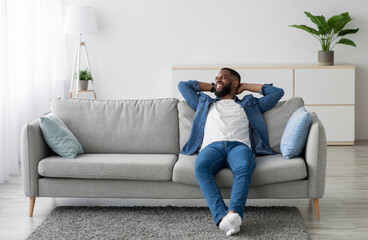 Cheerful attractive millennial african american male with beard sitting on couch and look at empty...