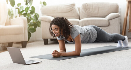Fototapeta premium Training At Home. Fit black woman doing yoga plank while watching online tutorial on laptop, exercising in living room, free space, panorama
