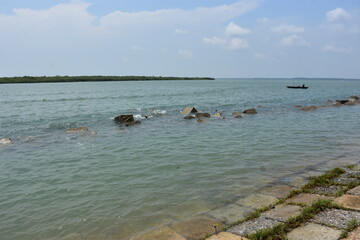 Fototapeta premium Calm Waters of Bakkhali River with Submerged Rocks and Stone-Paved Embankment, Framed by Distant Greenery and Gentle Waves in Cox’s Bazar, Bangladesh