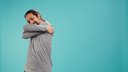Portrait of man standing in studio, hugging himself in gesture of comfort and self love, isolated...