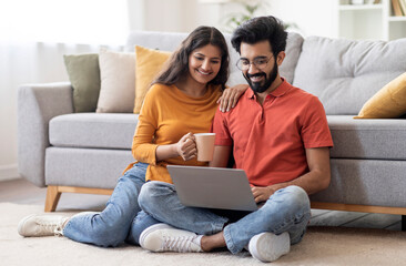 Smiling Indian Couple With Laptop And Coffee Relaxing In Living Room At Home, Happy Young Eastern...