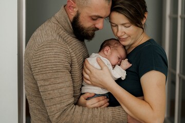 A couple lovingly holds their newborn baby in a cozy indoor setting. The mother gazes at the baby while the father supports them, showcasing a moment of tenderness and family bonding.