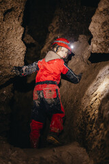 A 6-year-old girl in a protective suit and helmet with a flashlight in the middle of a natural underground cave is walking the tourist route of a caver