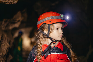 A 6-year-old girl in a protective suit and helmet with a flashlight in the middle of a natural underground cave is walking the tourist route of a caver