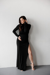 Naklejka premium Pregnant woman in a black dress posing against a white wall, smiling and holding her belly.