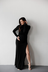 Fototapeta premium Pregnant woman in a black dress posing against a white wall, smiling and holding her belly.