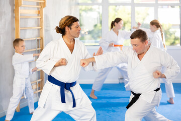 In gym, Man father in kimono learns to fight and, paired with male teacher, practices striking. Family karate coach conducts classes for parents and children