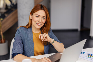 Portrait of smiling businesswoman sitting at corporate office with laptop and looking at camera.