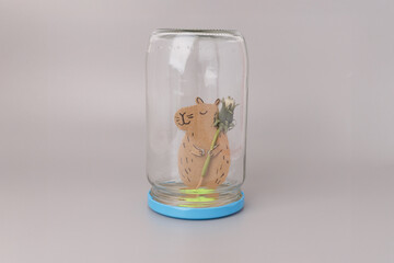 Upside-down glass jar on blue base with handmade brown paper capybara holding flower bud, ideal for DIY craft visuals, eco-friendly brands, kids education, storytelling, creative conten