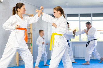 Fototapeta premium Young girl and woman in kimono and colored belt practicing karate punch block during group martial arts lesson in gym