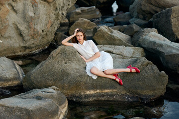 Elegant woman in white dress sitting on rock in water with vibrant red shoes