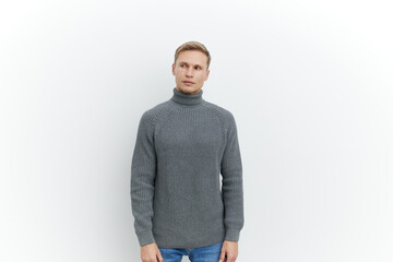 Fototapeta premium Full body portrait of a young Caucasian man with blonde hair wearing a gray knitted turtleneck sweater, looking at camera, standing against plain white background, studio shot, casual fashion, men