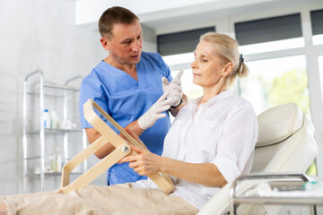 Positive elderly woman looking at mirror while skilled male cosmetologist showing results of hardware facial procedure in medical esthetic office