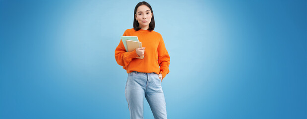 Young asian student with notebooks, standing in orange sweatshirt, looking at camera, posing over...