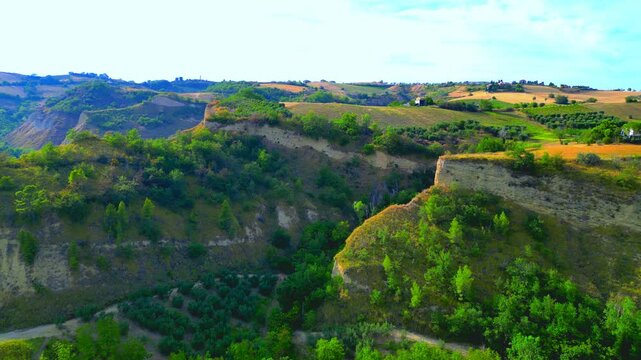 Momentous aerial push-in shot from Offida, a view near Chiesa di Santa Maria della Rocca with steep promontories topped by vegetation, valleys with trees, fields, thickets, sunlight imbuing the scene